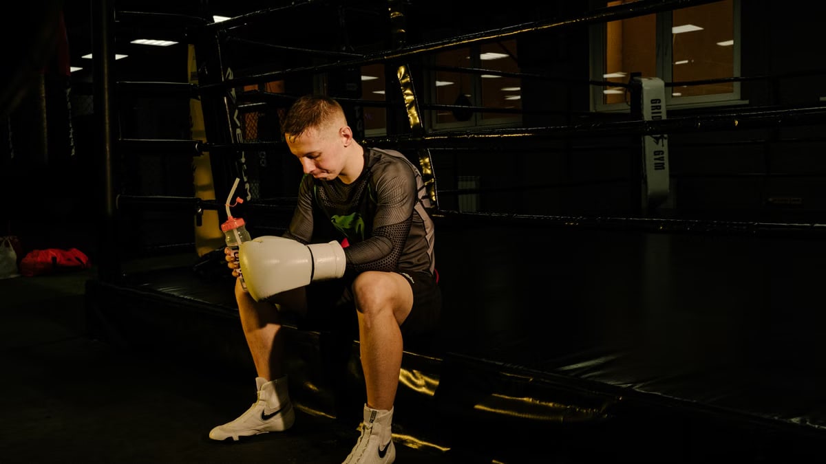 Young male boxer sitting in the ring holding gloves and a water bottle, taking a break.